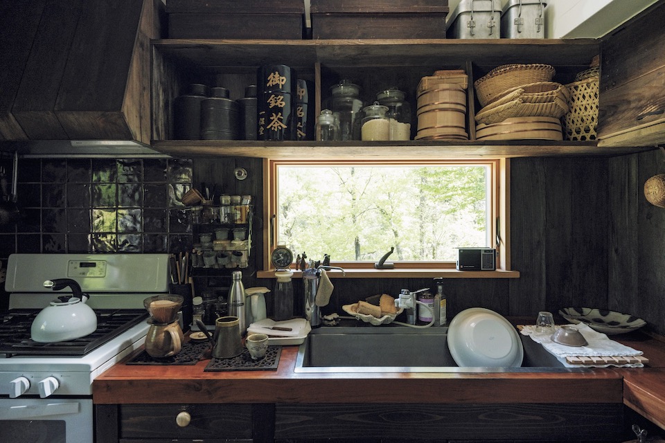 栃木県益子町にある『Loveit』店主のはったえいこさんの台所。窓からは四季折々の景色が楽しめる。森をイメージし、台所は茶系でまとめた。上 の棚には、愛飲するお茶の缶、米、小麦粉などの ほか、おひつやザルなど毎日使うものを収納。