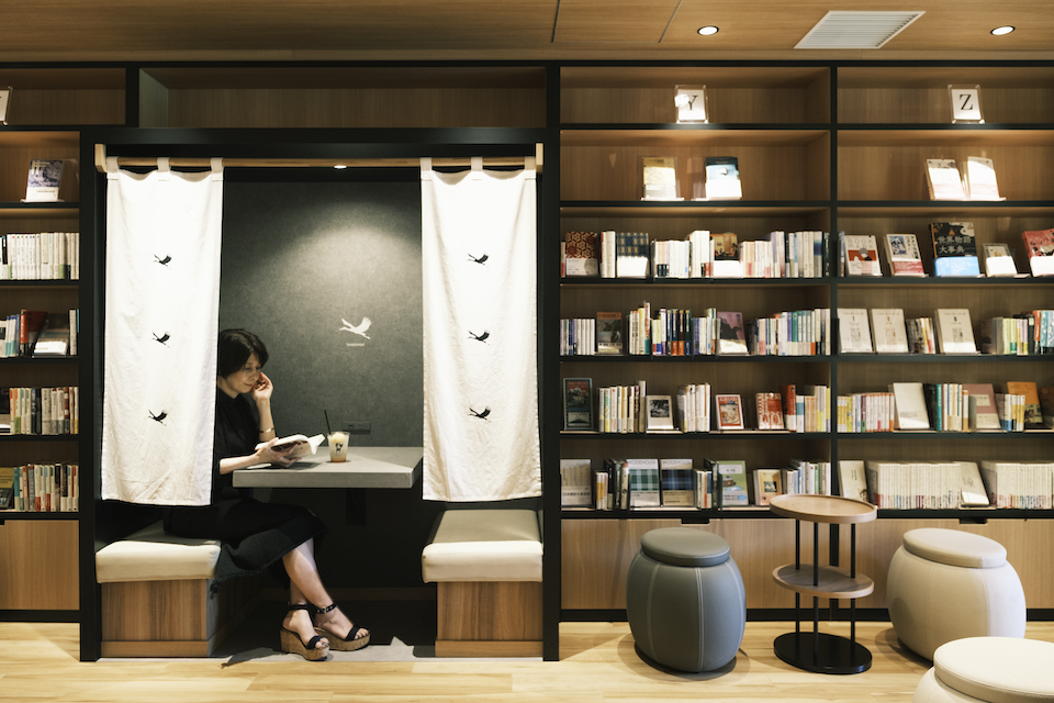 『短編喫茶Un』の2階、半個室のスペースで本を読むひうらさん。暖簾にはコウノトリの絵柄が。