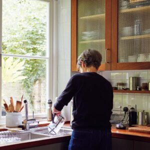 Elegance Margaret Howell 〈マーガレット・ハウエル〉に宿るエレガンス MHL アンドプレミアムオンラインストア | キッチンに立つマーガレット・ハウエル。庭に面した位置にあるキッチンには、自然光がたっぷり降り注ぐ。棚には日本で購入した茶器やオブジェも。