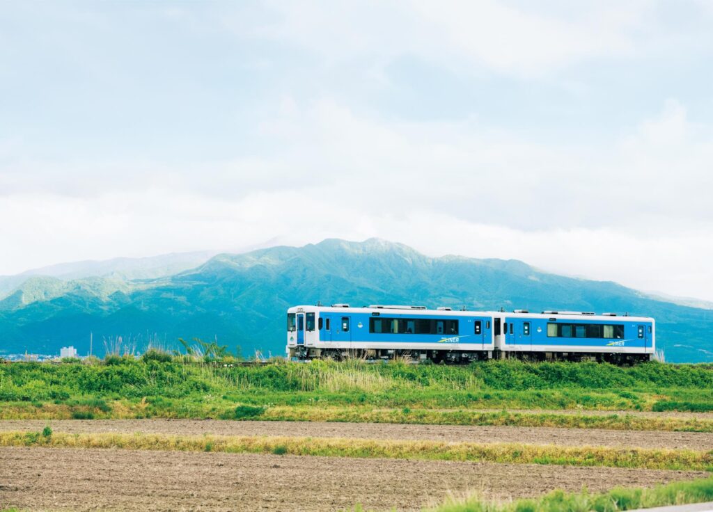 蔵王、月山、朝日連峰などの麗しい山々を遠くに望み、近くには里山。緑に染まる風景が囲む町には、明治期の山形県令・三島通庸の近代化構想に端を発する洋風建築が立ち並ぶ。
背景の蔵王に映える水色の車体はJR左沢線。山形市と山辺町の間にて。