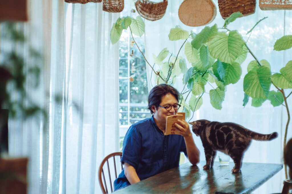 夜の古書店で、未知の本と出合う。歌人・穂村 弘さんの暮らしの本棚と、読書の時間の見つけ方。 | 自宅のリビングで読書をする穂村弘さん。愛猫のひるねがその姿を見つめる。