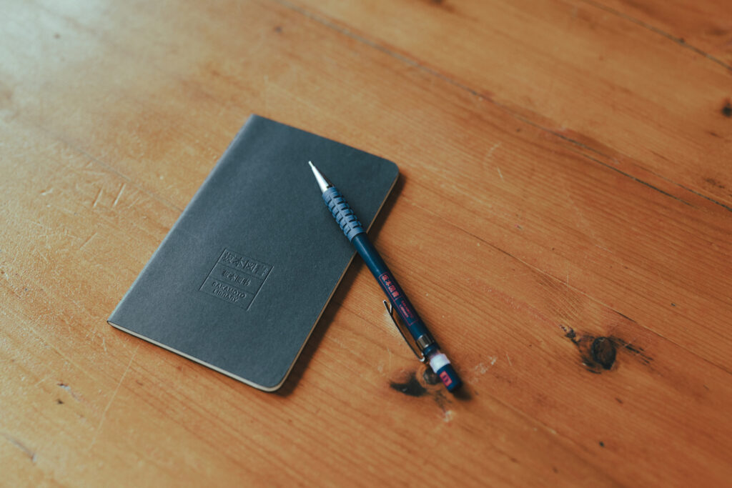 坂本龍一さんが残した本をきっかけに、新たな出合いを届ける。『坂本図書』と〈YAECA〉による図書空間『坂本ヤエカ』がスタート。 | 『坂本図書』の書籍やオリジナルグッズも販売。写真は、坂本さんがいつもポケットに入れていたノートとペンに、「坂本図書」の文字をあしらった。