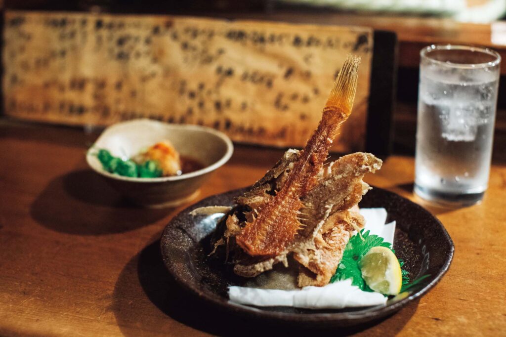 人を引き寄せる、ゆるやかな水辺のリズム。京都・鴨川、ローカルガイド。後編 | 骨までカリカリに揚げられたカレイのから揚げ（￥1,000）と芋焼酎「さつま白波」のソーダ割（￥700）の組み合わせが、『赤垣屋』でのお決まりという酒向さん。「ナスにしん
など、京都の味を街の先輩方に教えてもらったのもこの店」