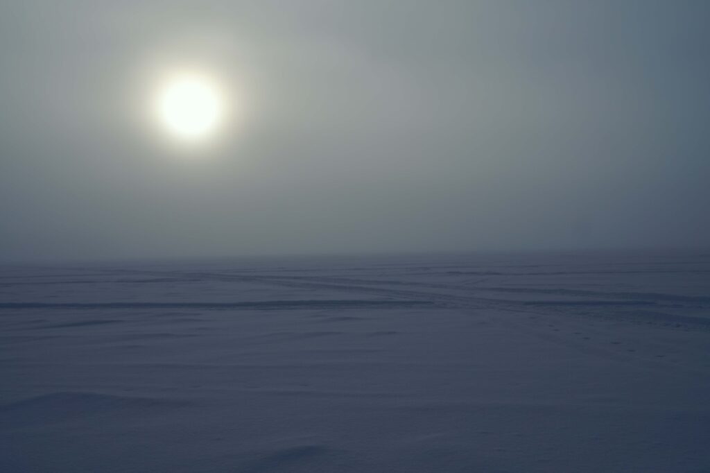 淡く差し込む冬の太陽が雪原に浮かび上がり、周囲は幻想的な光景が広がる。 | 凍った湖の雪原に淡く差し込む冬の太陽が浮かび上がり、幻想的な光景が広がる。