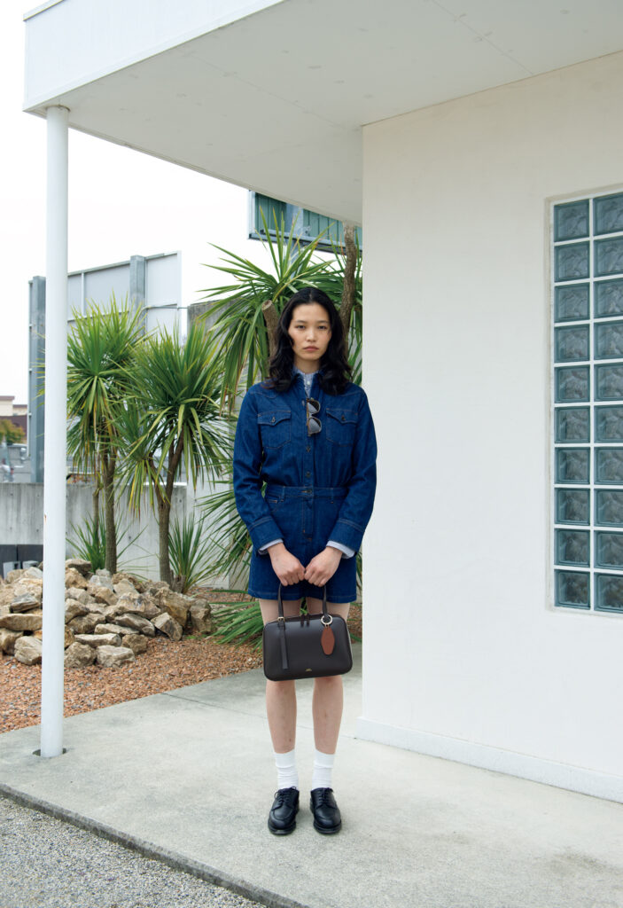 〈A.P.C.〉で人気のバッグ「Virginie」のスクエア型はコンパクトなボディが魅力。ハンドルと着脱可能なストラップで、ハンドキャリーも肩掛けも自在。深みのあるブラウンカラーの上質なスムースレザーが装いに洗練された気品を添える。バッグ￥99,000、チャーム￥14,300、デニムワンピース￥55,000、シャツ￥38,500、靴下￥4,950、サングラス￥40,700 (以上A.P.C.／アー・ペー・セー カスタマーサービス) 