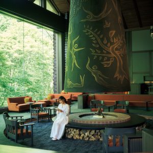 『奥入瀬渓流ホテル』で過ごす、川のせせらぎに包まれる癒やしの滞在。坂本美雨さんと巡る、青森への旅。ロビー中央に擁する岡本太郎作の大暖炉《森の神話》に腰かけ、本を開 く坂本さん。岡本太郎に関する本が並ぶ本棚もあり、外の景色を眺めながらゆったり過ごせる。 | ロビー中央に擁する岡本太郎作の大暖炉《森の神話》に腰かけ、本を開く坂本さん。岡本太郎に関する本が並ぶ本棚もあり、外の景色を眺めながらゆったり過ごせる。