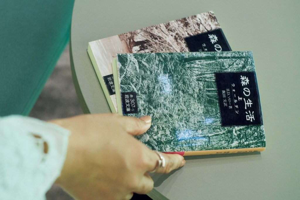 自然にまつわる本が並ぶ「渓流‒BASE」で坂本さんが手に取ったのは、H.D.ソロー著、飯田実訳による『森の生活』。『奥入瀬渓流ホテル』で過ごす、川のせせらぎに包まれる癒やしの滞在。坂本美雨さんと巡る、青森への旅。ロビー中央に擁する岡本太郎作の大暖炉《森の神話》に腰かけ、本を開 く坂本さん。岡本太郎に関する本が並ぶ本棚もあり、外の景色を眺めながらゆったり過ごせる。 | 自然にまつわる本が並ぶ「渓流‒BASE」で坂本さんが手に取ったのは、H.D.ソロー著、飯田実訳による『森の生活』。