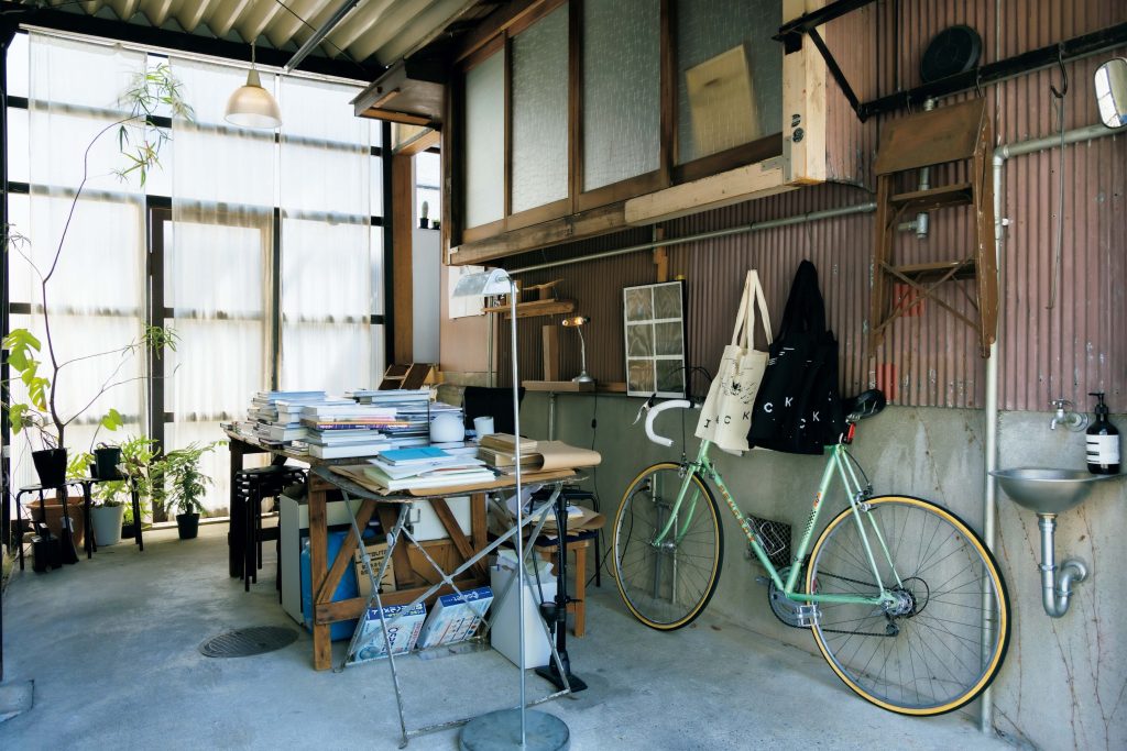 建築やアートを巡る旅。石川・金沢で出合った17のスポット。中編 IACK  ワインショップ横の小さな入り口から、路地を入った先に、主に写真集を扱うブックショップ＆ギャラリー『IACK（アイアック）』（金沢市高岡町18‒3）がある。1階では写真家である店主・河野幸人さんが、ガレージだった場所を活用し、アートブックフェアのブースのように一台のテーブルの上に本をどさりと並べている。「家と家の間の細い隙間を抜けていくアプローチ。秘密の場所に来たようなワクワク感も味わえます」と、松本さん。 | IACK
ワインショップ横の小さな入り口から、路地を入った先に、主に写真集を扱うブックショップ＆ギャラリー『IACK（アイアック）』（金沢市高岡町18‒3）がある。1階では写真家である店主・河野幸人さんが、ガレージだった場所を活用し、アートブックフェアのブースのように一台のテーブルの上に本をどさりと並べている。「家と家の間の細い隙間を抜けていくアプローチ。秘密の場所に来たようなワクワク感も味わえます」と、松本さん。