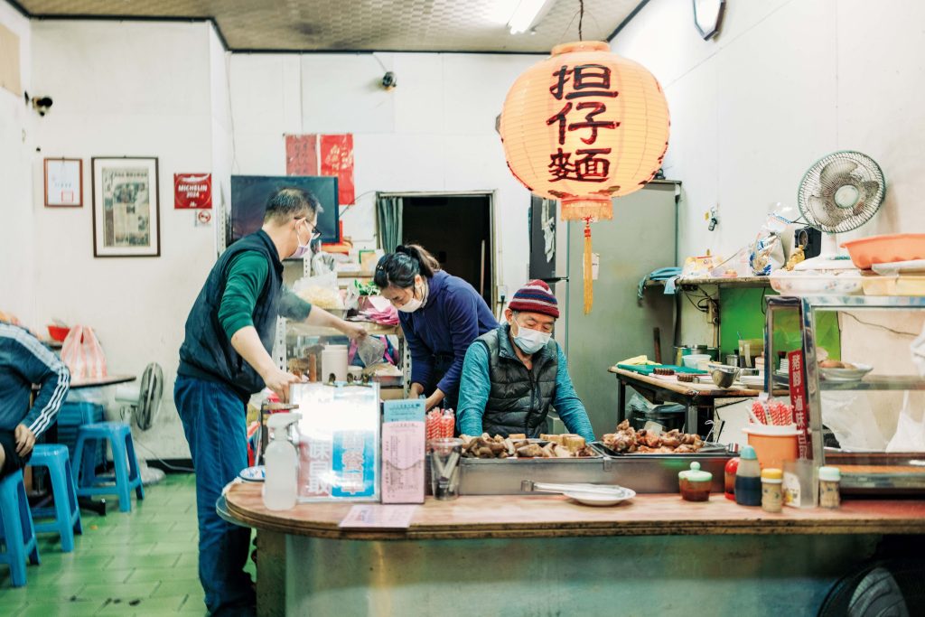 老若男女に愛される、地元ならではの味。台南フードを味わう。族経営。「ロータリーのそばにあるので、バイクや行き交う人など、街の風景も一緒に楽しめます。 | 伝統的な座卓調理台で麺づくりに勤しむ2代目店主。3代目の息子も含めた家族経営。「ロータリーのそばにあるので、バイクや行き交う人など、街の風景も一緒に楽しめます。