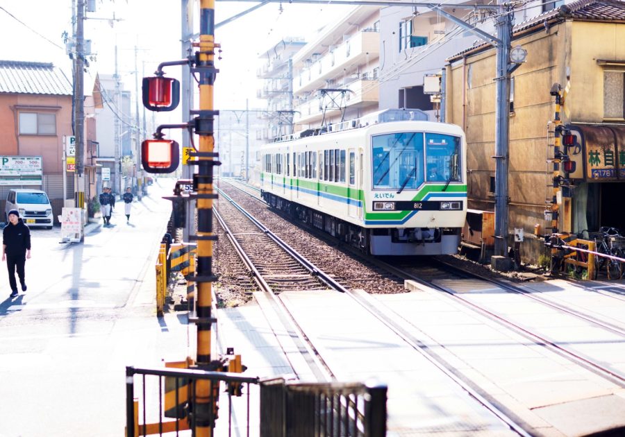 京都・一乗寺の〝えいでん〞こと叡山電鉄 | 鴨川デルタに程近い南側の始発駅・出町柳から、鞍馬、八瀬比叡山口といった山間部まで、左京区を南北に繋ぐ市民の足〝えいでん〞こと叡山電鉄は、昨年開業100年を迎えた。出町柳から乗車しておよそ5分、3つ目の駅が一乗寺だ。写真の2両編成、800系（デオ810形）の帯色は沿線の山並みをイメージした緑。無人駅の小さなホームからは、街のメインストリートである曼殊院道の商店街
を横切る車両を近距離で眺められる。「乗るたびにちょっとだけ旅行の気分になります」とは案内人の日髙さん。ちなみに「一乗寺」の地名の由来となったのは同名の寺院。紫式部が仕えたとして知られる藤原彰子によって建立されたとされるが、焼き討ち、再建を経て南北朝の戦乱で消失。以来、廃絶した。